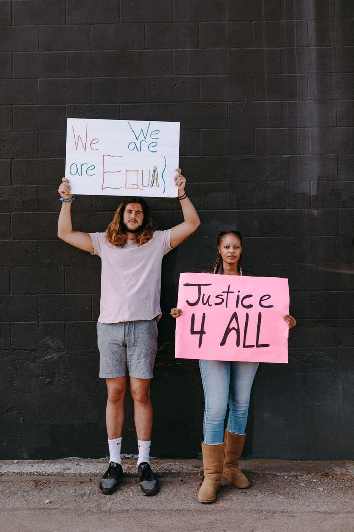 Two activists holding signs advocating for equality and justice against a dark urban wall.