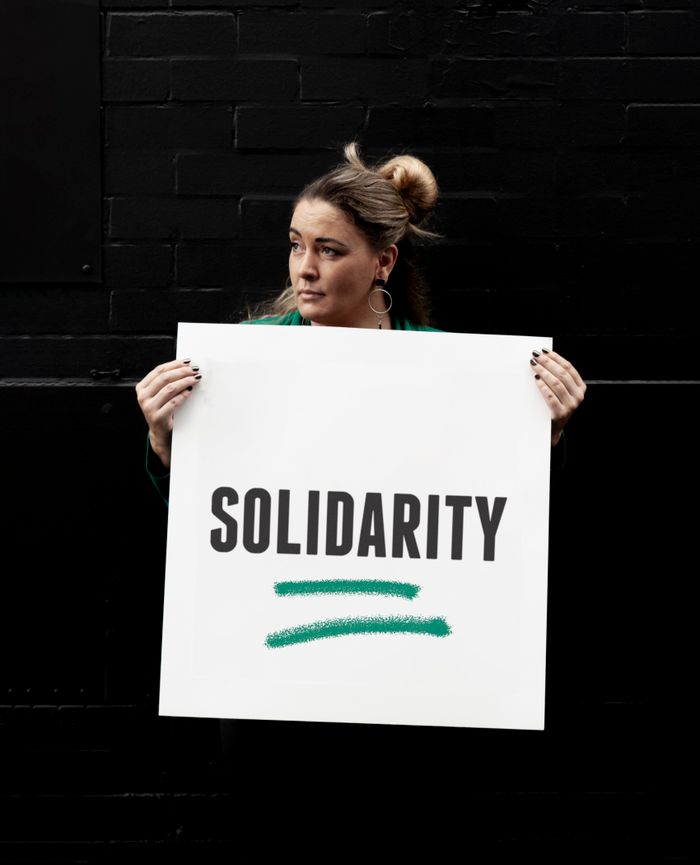 our-story Adult female showing banner with Solidarity inscription and looking away against black wall on city street